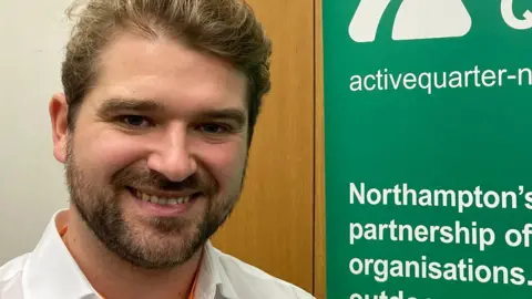 A man with short brown hair and a beard smiles at the camera. He is wearing a white collared shirt and an orange lanyard.