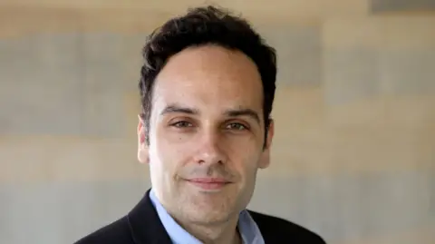 Dr Javier Andreu-Perez is smiling. He has black hair and stubble. He is wearing a black suit jacket and a pale blue shirt. There is a wall behind him, although it has been blurred.
