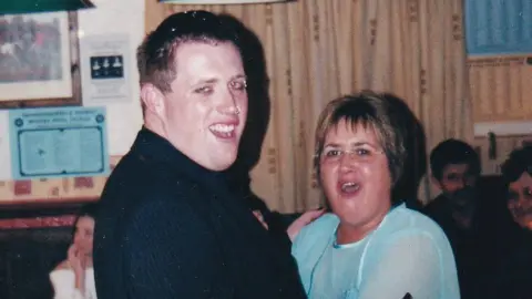 Photograph of Dale Morgan (left) with Judith Rhead(right). Judith has short brown hair with blonde highlights. She wears a blue sheer long sleeve top, thin rimmed reading glasses and smiles at the camera. Dale wears a black pinstripe shirt, a black waistcoat and a nose ring. He has brown hair and wears black eyeliner. 