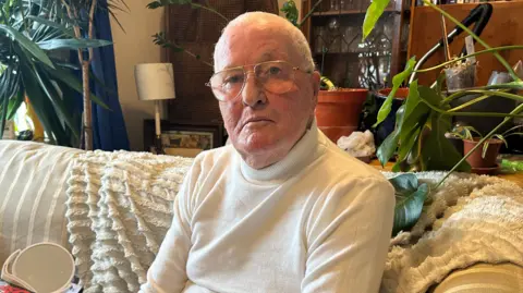 BBC Timothy Willis is pictured sitting on a striped sofa which is cream in colour. There is a fluffy cream blanket draped over the sofa and a sideboard and plants behind it. Mr Willis, who is an older man, is wearing a cream roll-neck jumper and glasses and he has white hair. He has a serious expression and is looking directly at the camera.