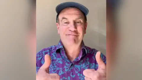 Marc Williamson/YouTube Mr Williamson has both his thumps up as he smiles for the camera. He is wearing a blue and purple shirt and a blue flat cap.