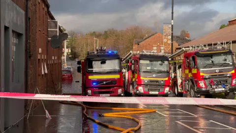 Fire appliances parked three abreast in Kings Row, Coalisland, on Saturday. The red-brick street beyond is flooded with water reaching just below the rear number plate of red car.  Hoses are lying on the ground and the street is taped off. 