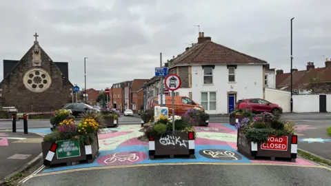 A row of planters blocking the end of a road, with space for bicycles to pass between them. One of the planters has a "road closed" sign on it. On the other side is a queue of traffic heading along a main road.
