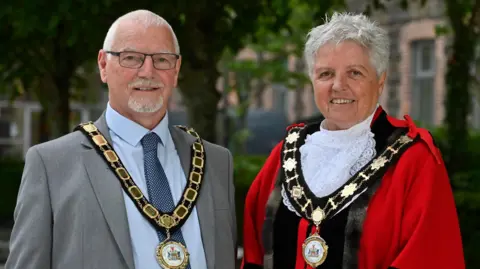 Antrim and Newtownabbey Borough Council Henry Cushinan and Alison Bennington are standing next to each other. Cushinan is wearing a grey suit and Bennington is wearing official red robes. Both have gold chains of office on. 