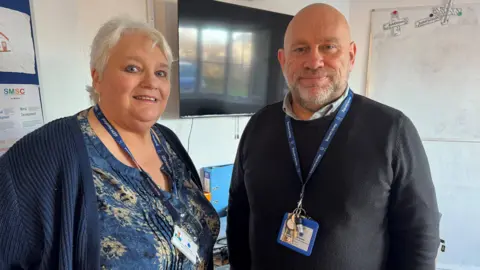 A woman wearing a blue jumper and blue flowery shirt, stood alongside a man wearing a black jumper. They are both wearing lanyards, and are stood in a school setting.