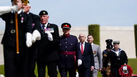 PA Media A parade of people in military uniform, with a man wearing a navy blue suit in the middle. There is a man in a navy uniform carrying a red poppy wreath, while the person at the front of the parade is carrying a flag.