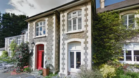 A picture of the outside of the hotel. The building is all brick, grey and white, and has a red door. There is plants surrounding the hotel with hedges next to it.