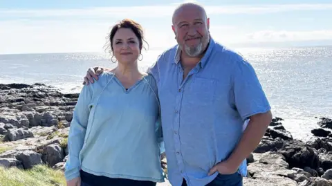 Actors Ruth Jones and Steve Speris standing with the sea and shelving rocks behind them. 