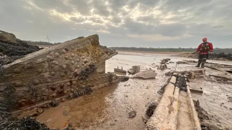 Rusting metal scraps of the Severn Railway Bridge are strewn across the vast muddy riverbed of the Severn. A man in red rescue gear stands surveying the debris.