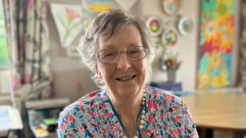 Alice Cunningham/BBC An elderly lady with short grey hair smiles at the camera as she sits in a communal living area. She wears a blouse with a blue, red and black pattern on it. She is also wearing a necklace made of gold pearls.