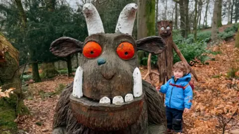 A wooden sculpture of the Gruffalo - which is a brown furry creature with large white horns and a wart at the end of its nose - stands on a forest path. There is another sculpture in the background as well as a young boy admiring it.