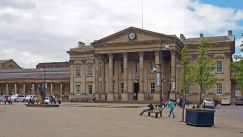 Getty Images A photo of the outside of Huddersfield station. There is a large pedestrianised area in front of the Victorian building. A number of people are milling around and there are a few taxis parked at either side of the building.