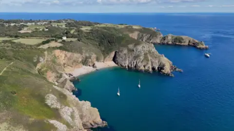 BBC An aerial image of the island of Sark, showing a beach and cliff faces under sunny skies. There re a number of boats bobbing in the sea.