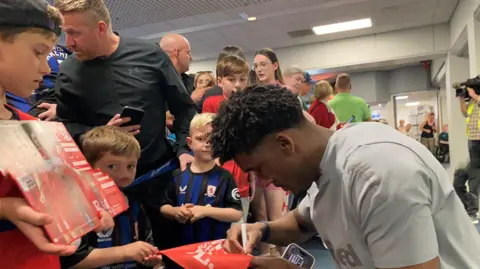 A team member is crouched in front of a deep crowd at the airport and is signing a red flag for a young fan, while other youngsters in both blue and red Middlesbrough kits look on. The player has short, curly dark hair and is wearing a grey shirt.