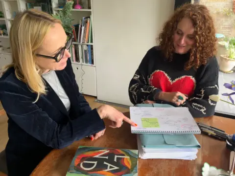 BBC/Katie Razzall Katie Razzall on the left, blonde hair, glasses in dark blue velvet jacket seated at a table, pointing to note book. Maggie O'Farrell, auburn curly hair in dark jumper with red heart in the middle.
Book shelves behind them on left Door to garden with plants in pots on the right