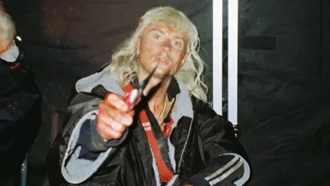 Sam Locke Locke wears a shoulder length blond wig. He has dirt and blood on his face. He holds a pocket knife to the camera. 