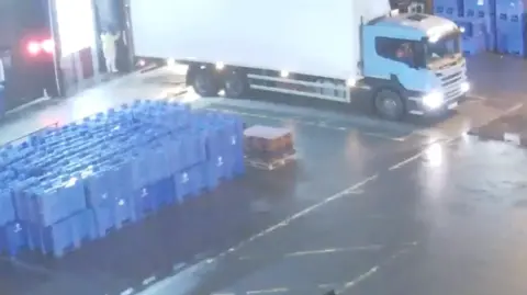 Health and Safety Executive A stack of blue crates two high and a white lorry in a loading bay with a man in a white outfit at night.