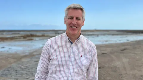 Helier Du Veulle standing on a beach at low tide. He has short grey hair and is wearing a blue and red check shirt. 