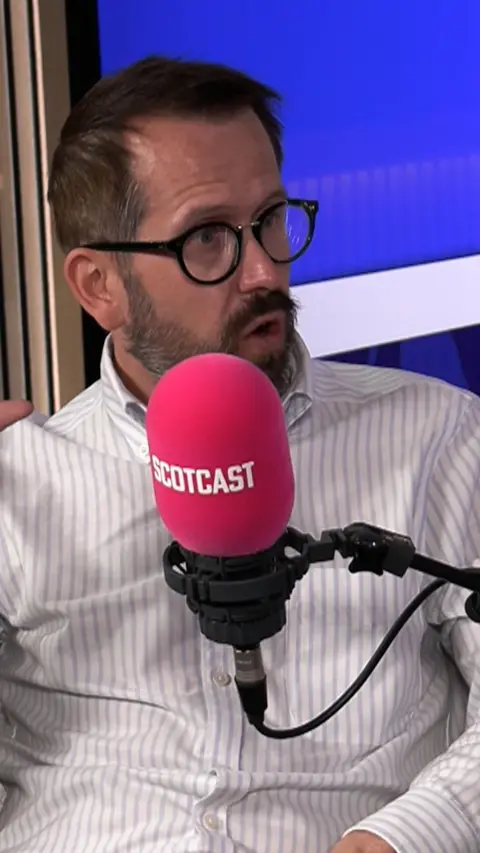 Man in white shirt with beard and glasses in front of red microphone