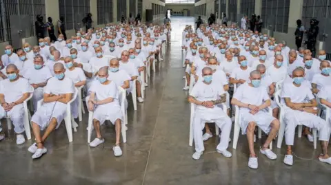 A group of hundreds of men with shaved heads, wearing white t-shirts and shorts or trousers and blue surgical masks, are seen sitting on white plastic chairs in an open space at a confinement centre in El Salvador