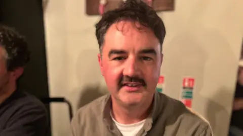 A man with black hair and a black moustache stares at the camera. He has a brown shirt on and a white t-shirt underneath. Behind him you can see a plain coloured wall and to his left is a partial view of a man in a blue jumper with black hair. 