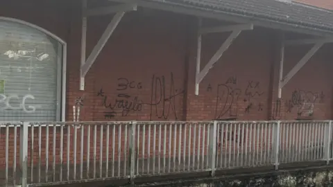 LDRS The image shows a red brick wall surrounded by a grubby white railing. Paint is peeling from white rafters. The walls have graffiti tags scrawled across them, in black. One tag appears to read "waylo", another says "3CG", while others are indecipherable. 