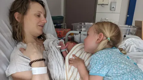 Louise in a hospital bed with her newborn baby and older daughter, who is wearing a blue floral dress and has blonde hair