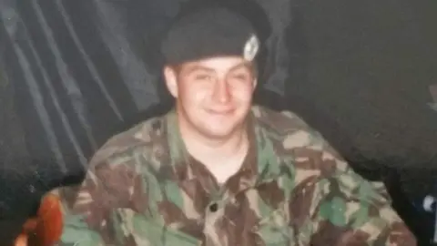 MIKE RYAN An old photo of a young Mike Ryan in an army uniform as he smiles in front of a black background.