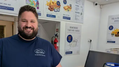 Seniors Dominic Horabin with brown hair and beard wearing a navy blue t-shirt with a logo saying 'Seniors' in a fish shape stands behind the counter. He is smiling. 