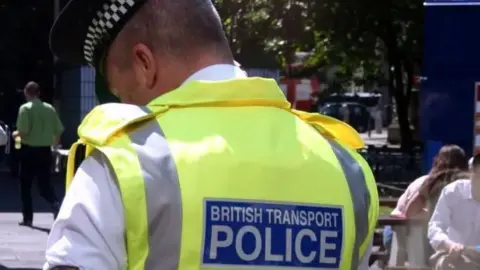 A British Transport Police officer wearing a yellow vest and white shirt. The back of the vest has 'British Transport Police' printed on it in white letters on a blue background.