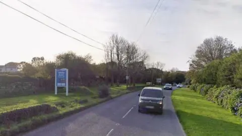 Google Google Street View of the A392 at White Acres near Newquay. Three vehicles are on the road heading towards the camera. A blue sign for White Acres Holiday Park is on a grass verge on the left-hand side. Trees and a stone wall line the road.