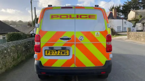 A stock image of a police van pictured from behind. The van is parked in a rural village.