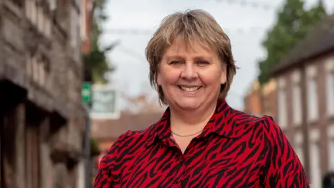 Shropshire Council Woman with short light brown hair is looking at the camera and smiling. She is wearing a red and black animal print blouse and a gold necklace. There is a blurred high street behind her.