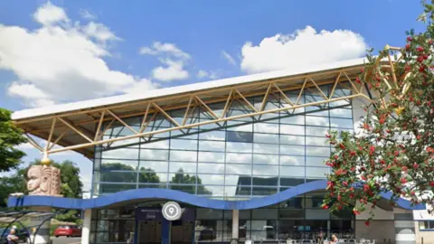 A glass-fronted building with a sloping roof stands against a blue sky.
The building has a blue canopy in a wave shape halfway down the front face of the building.