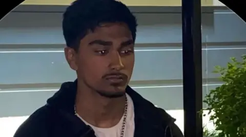 Young man, with dark hair and trimmed moustache poses looking away from camera. He wears a white t-shirt, chain necklace and dark hoodie. He stands in front of a window and a small green plant is positioned to the right of him.