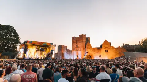 A large crowd is gathered in front of an outdoor concert stage, beside illuminated castle ruins at dusk.