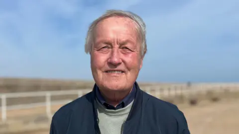 Bill is standing on a coastal promenade with hands behind their back, wearing a dark jacket and light jumper, with benches, railings, and open sky in the background