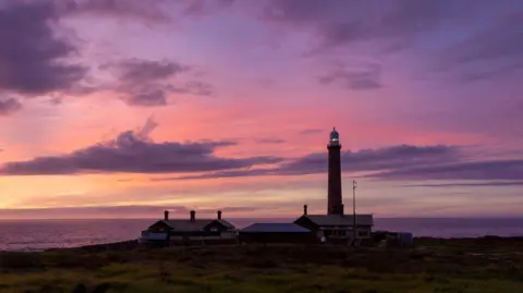 Nick Esser/Parks The Victoria Gabo Island lighthouse, a tall structure under a purple sunset sky, with the ocean again in the background.