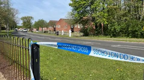 In the foreground, blue and white police tape has been attached to a black fence. In the background, forensics officers in white suits are inspecting ground near a fence with a row of houses behind.