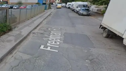 A general view of the road. It has potholes and damaged tarmac and has vehicles on the right-hand side and an industrial building on the left.