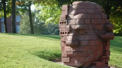 A brick-built statue depicting the head of Josiah Wedgwood in a stylised fashion. It is positioned on a grass verge.