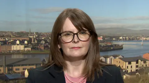 Caoimhe Archibald wears glasses. She has a pearl necklace on and is wearing a pink top under a black jacket. She has brown hair.