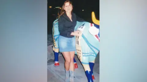 Gosia Nowak stands in front of a blue-coloured statue of a bull. Gosia is wearing a black shirt and a denim skirt with blue boots. She has brown hair below her shoulders and she is smiling at the camera