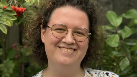 Emily Rhodes Emily Rhodes, with short curling dark hair haloed around her head, and round metal framed glasses, smiling at the camera with green shrubs with red berries behind her