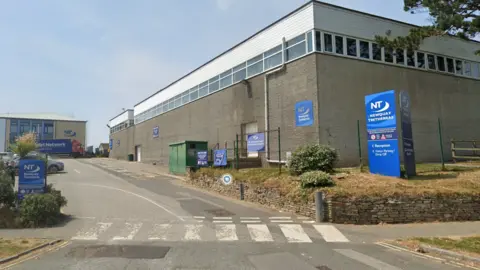 The gateway of the school. The entrance has a zebra crossing across it and there are many blue signs reading Newquay Tretherras. The nearest one points to reception, visitor parking and drop off. Beyond the entrance are large brick buildings with windows just below the roofline. Further back is a brick building with large windows. 