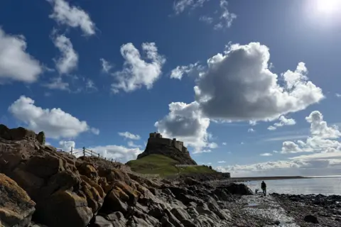 Toby the Gardener Grey clouds dot the sky with their edges lit by the rays of sunshine coming from the top right of the image. Below, there is a castle sitting atop a headland, with someone walking on the path leading up to it. There is a rocky shoreline in the foreground.