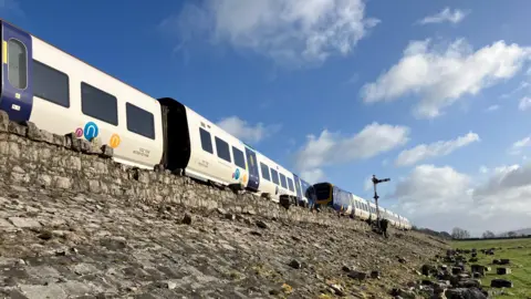 BBC Three carriages of the derailed train are visible on the tracks above a rocky embankment. The derailed section of the train is not visible