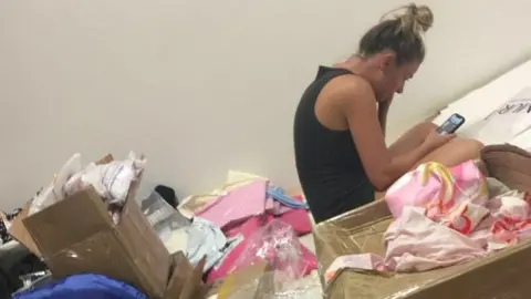 Olivia Prince Olivia hunched over her phone with her hair in a bun surrounded by boxes of clothes. 