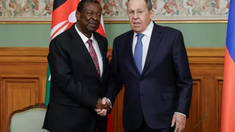 Reuters Russian Foreign Minister Sergei Lavrov (R), in a dark navy suit and light blue tie with white polka dots, shakes hands with Kenyan Foreign Minister Musalia Mudavadi (L), in a black suit with a maroon and pink paisley tie, during a press conference in Moscow - 16 March 2026. Behind Mudavadi can be seen the Kenya flag. To Lavrov's side the Russian flag can be partially seen.