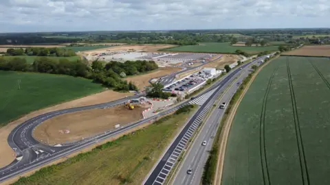 John Fairhall/BBC Aerial photo shows a park and ride site under construction, close to a dual carriageway road. The area is surrounded by green fields.
